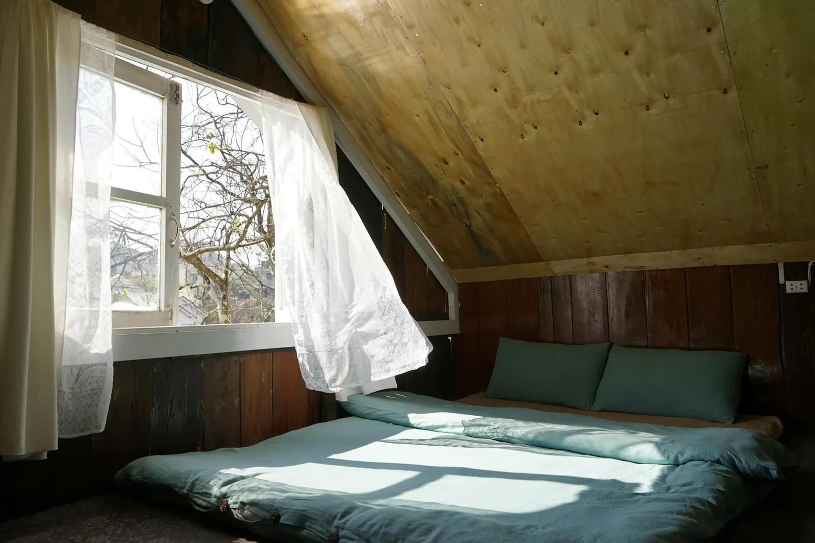 Sunlit attic bedroom with an open window and white curtains blowing inwards in the breeze