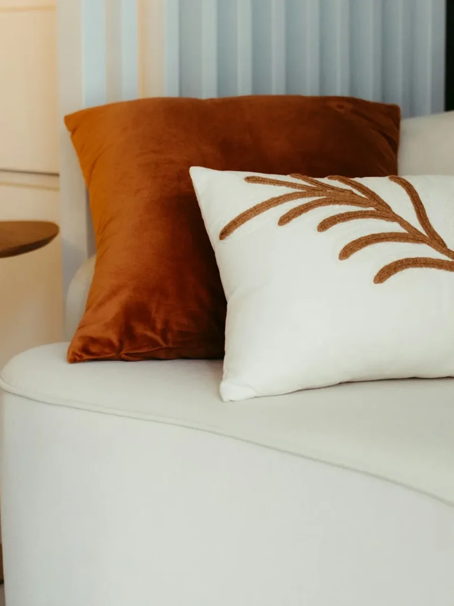 A cream bouclé sofa with a rust velvet cushion and an embroidered leaf cushion beside a brass side table