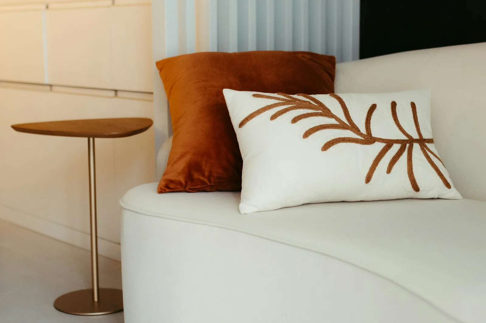 A cream bouclé sofa with a rust velvet cushion and an embroidered leaf cushion beside a brass side table