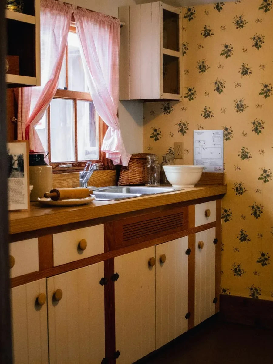 Small vintage kitchen with small-print floral wallpaper, cream cabinets and pink-curtained window
