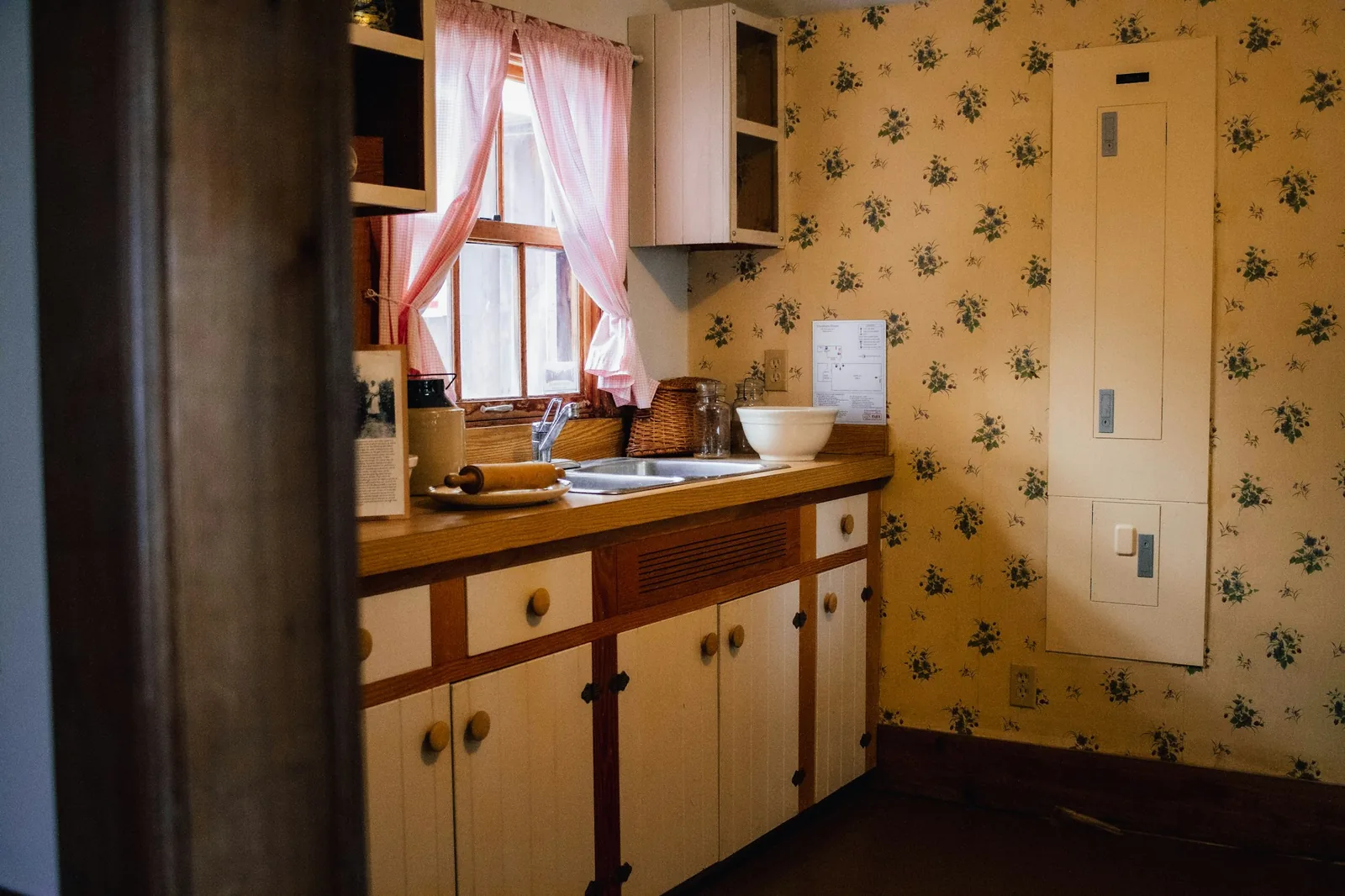 Small vintage kitchen with small-print floral wallpaper, cream cabinets and pink-curtained window
