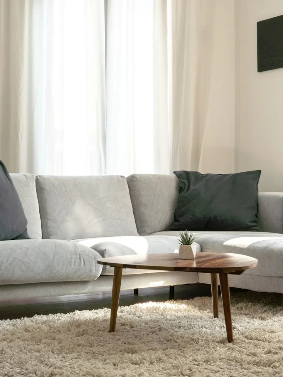 Light grey L-shaped sofa with chaise end, round wooden coffee table and shaggy rug by sheer curtains