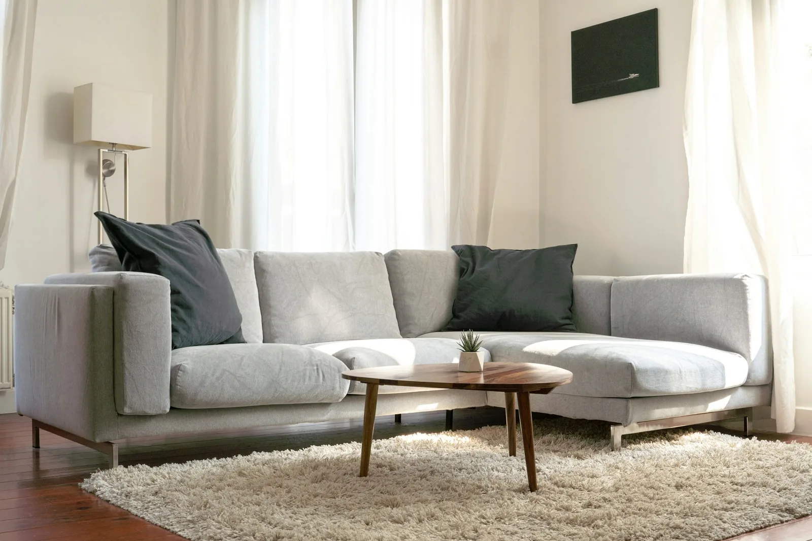 Light grey L-shaped sofa with chaise end, round wooden coffee table and shaggy rug by sheer curtains