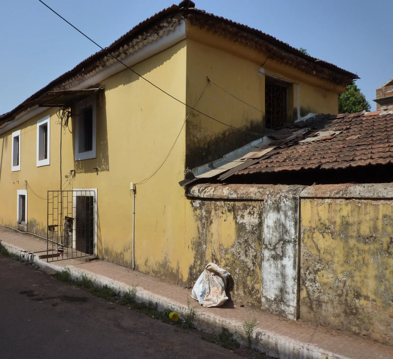 A yellow lime-washed Fontainhas house beside a weathered, patchy old plastered boundary wall and tiled roof