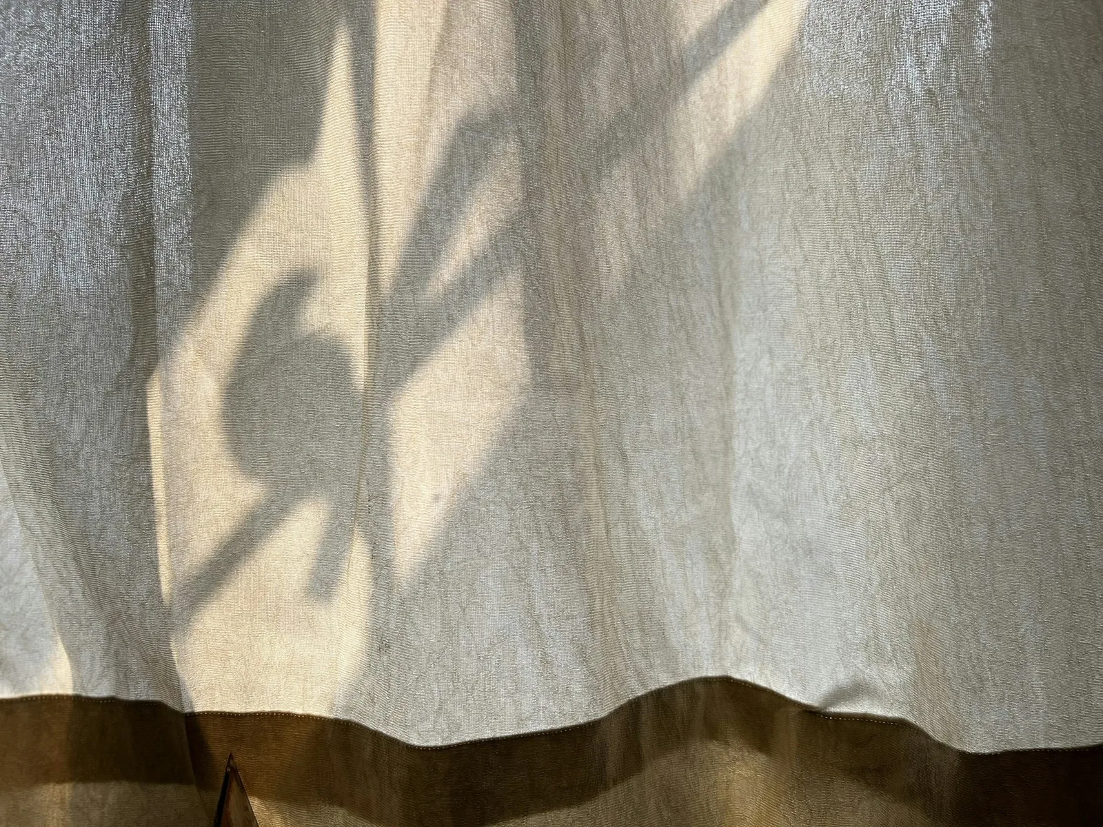 Sunlight casting a plant's shadow through a thin unlined curtain with a darker hem band