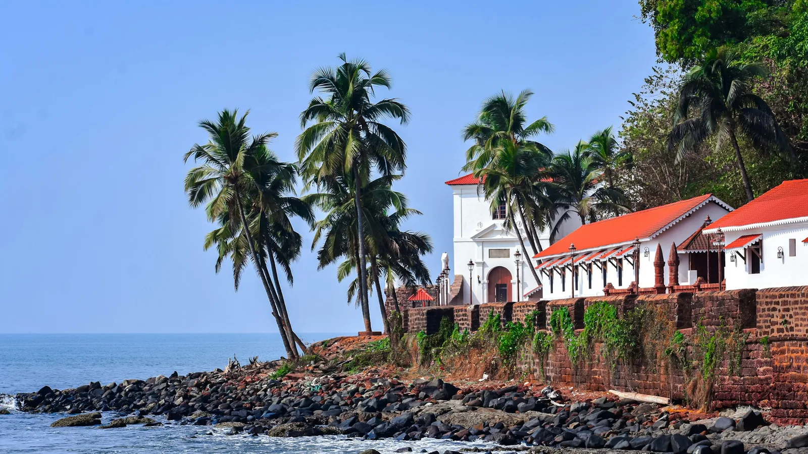 Whitewashed buildings with red-tiled roofs and coconut palms above a rocky stretch of tropical coastline