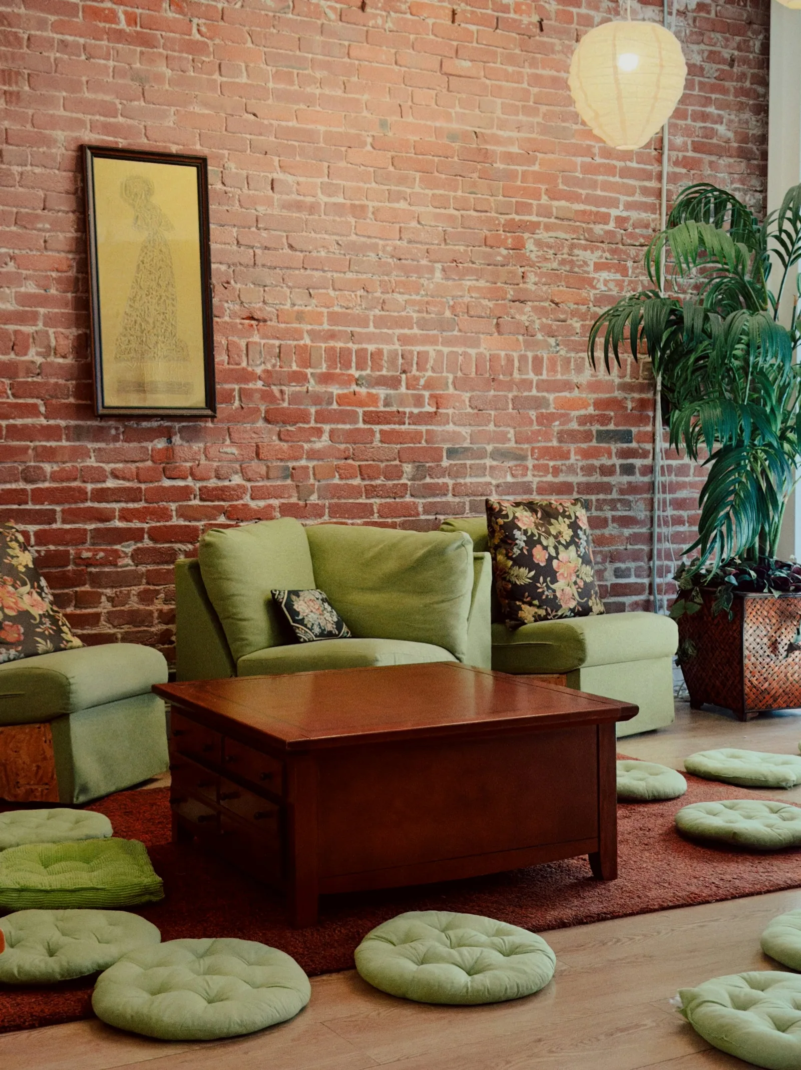 A brick-walled living room with green armchairs, patterned cushions, floor poufs and a paper lantern