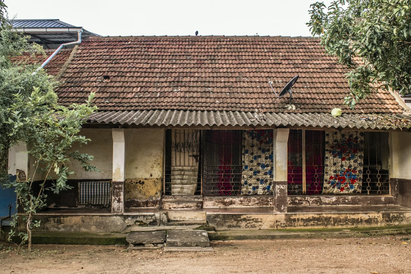 Weathered single-storey Indian house with a clay-tiled roof, grilled verandah and patterned cloth screens