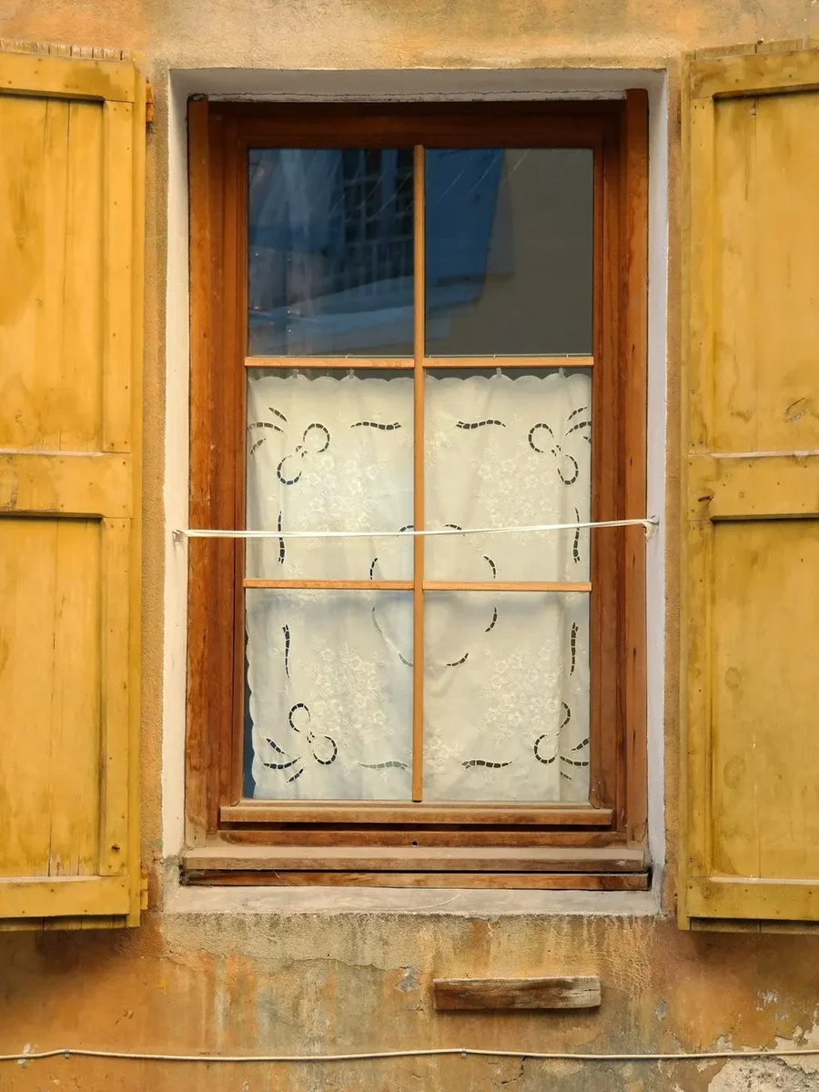 Small-paned wooden window with lace curtain and open yellow shutters on a weathered ochre wall