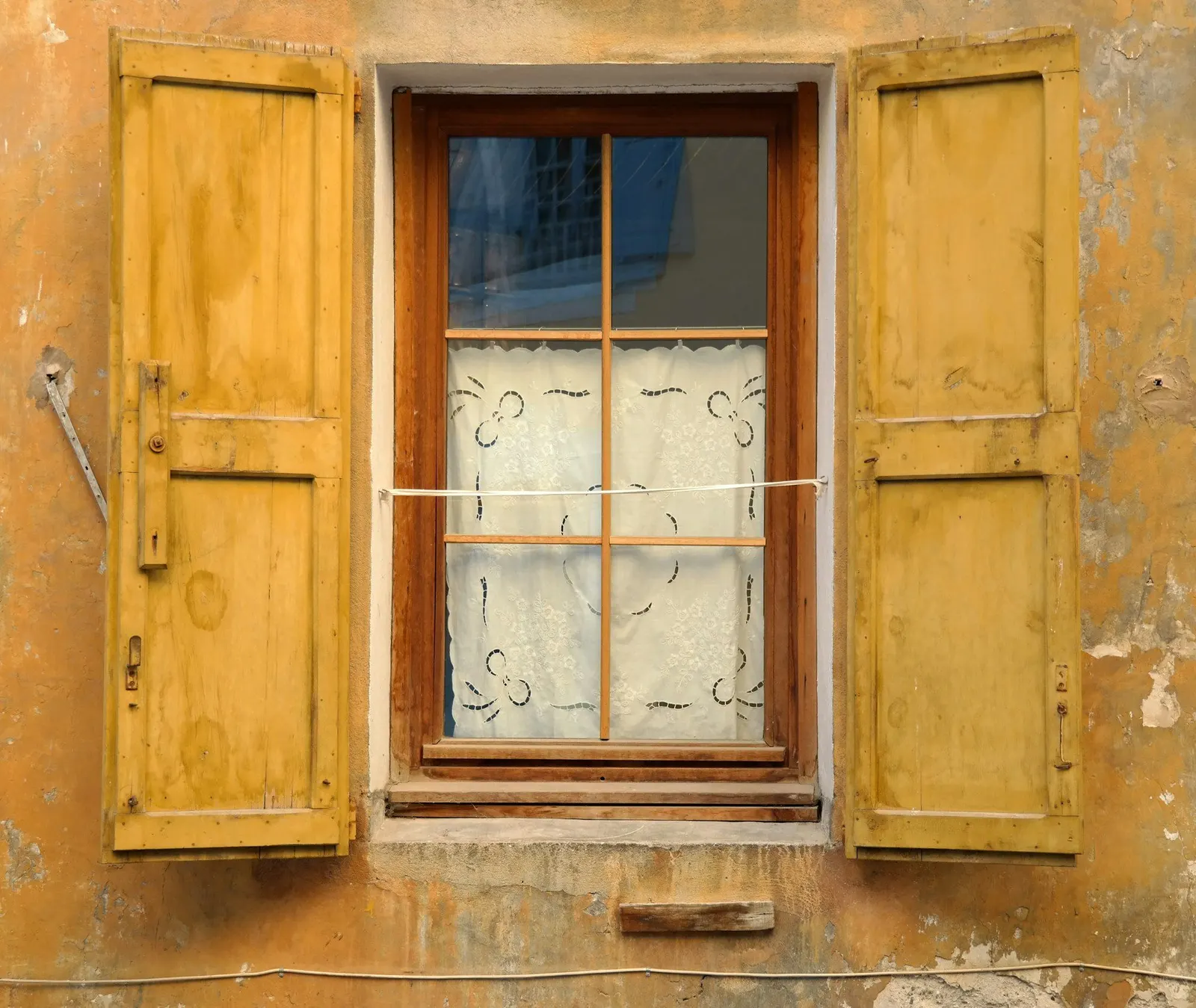 Small-paned wooden window with lace curtain and open yellow shutters on a weathered ochre wall