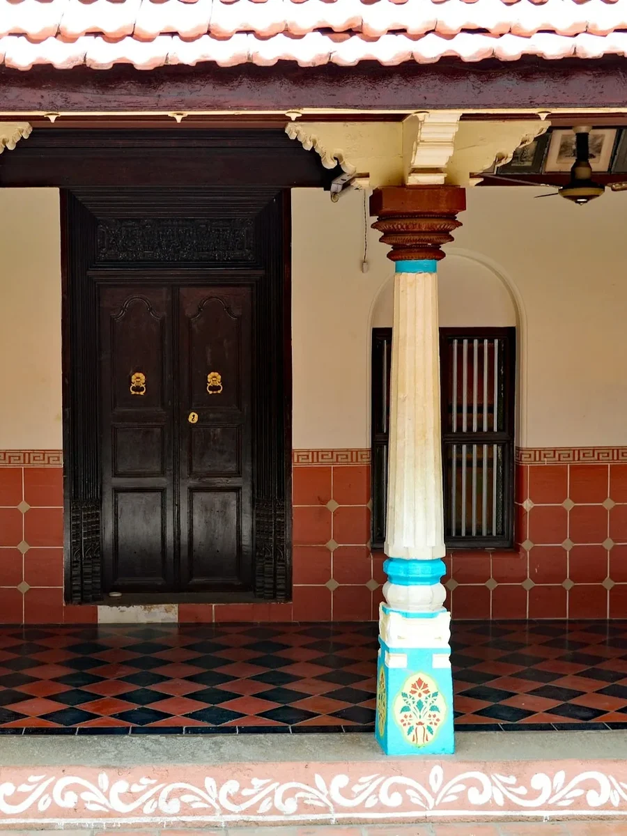 An Indian house verandah with a red and black diamond-pattern tiled floor, turned pillars and dark carved doors