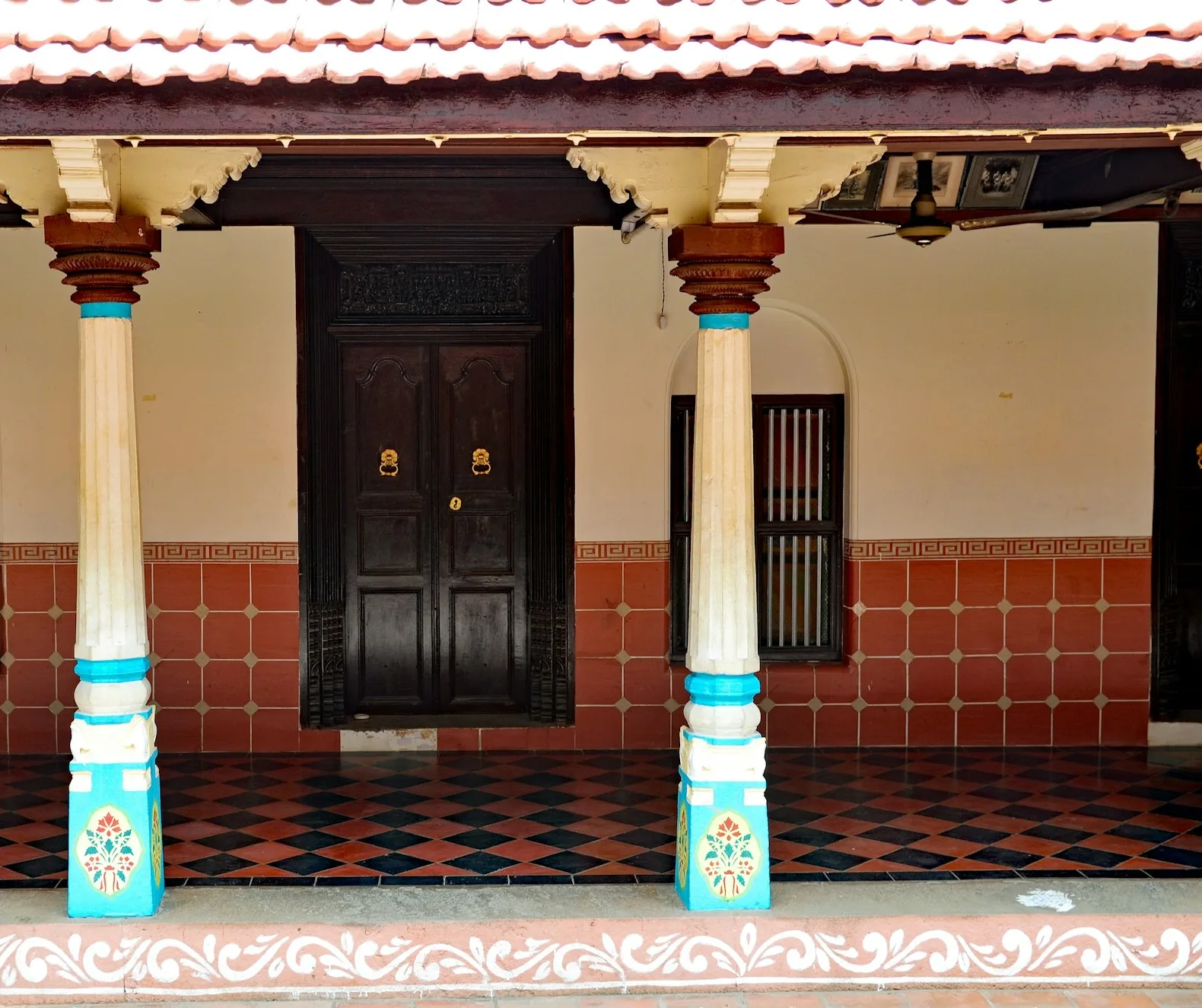 An Indian house verandah with a red and black diamond-pattern tiled floor, turned pillars and dark carved doors