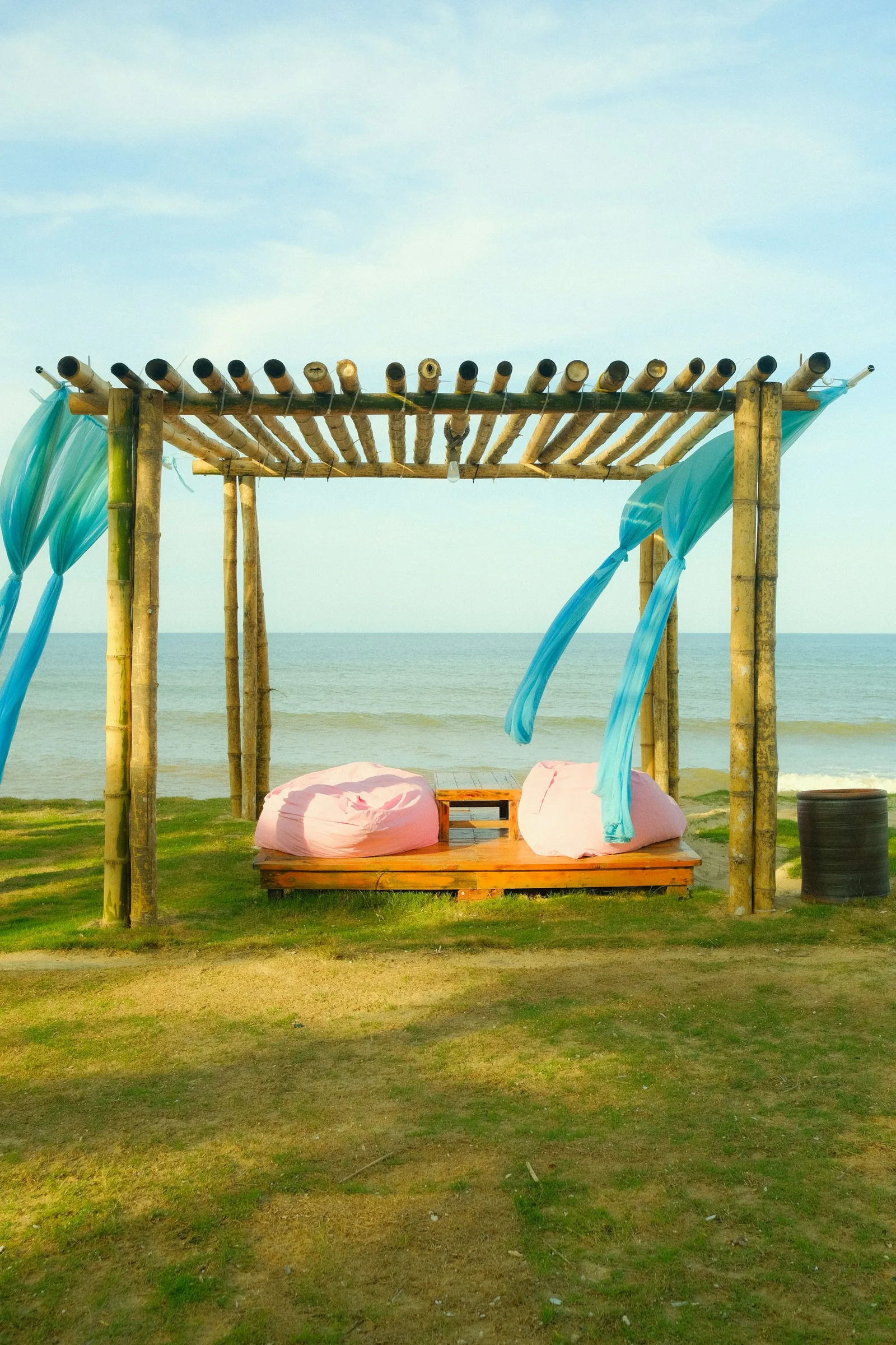A bamboo pergola draped with flowing turquoise fabric over a low daybed on a lawn facing the sea