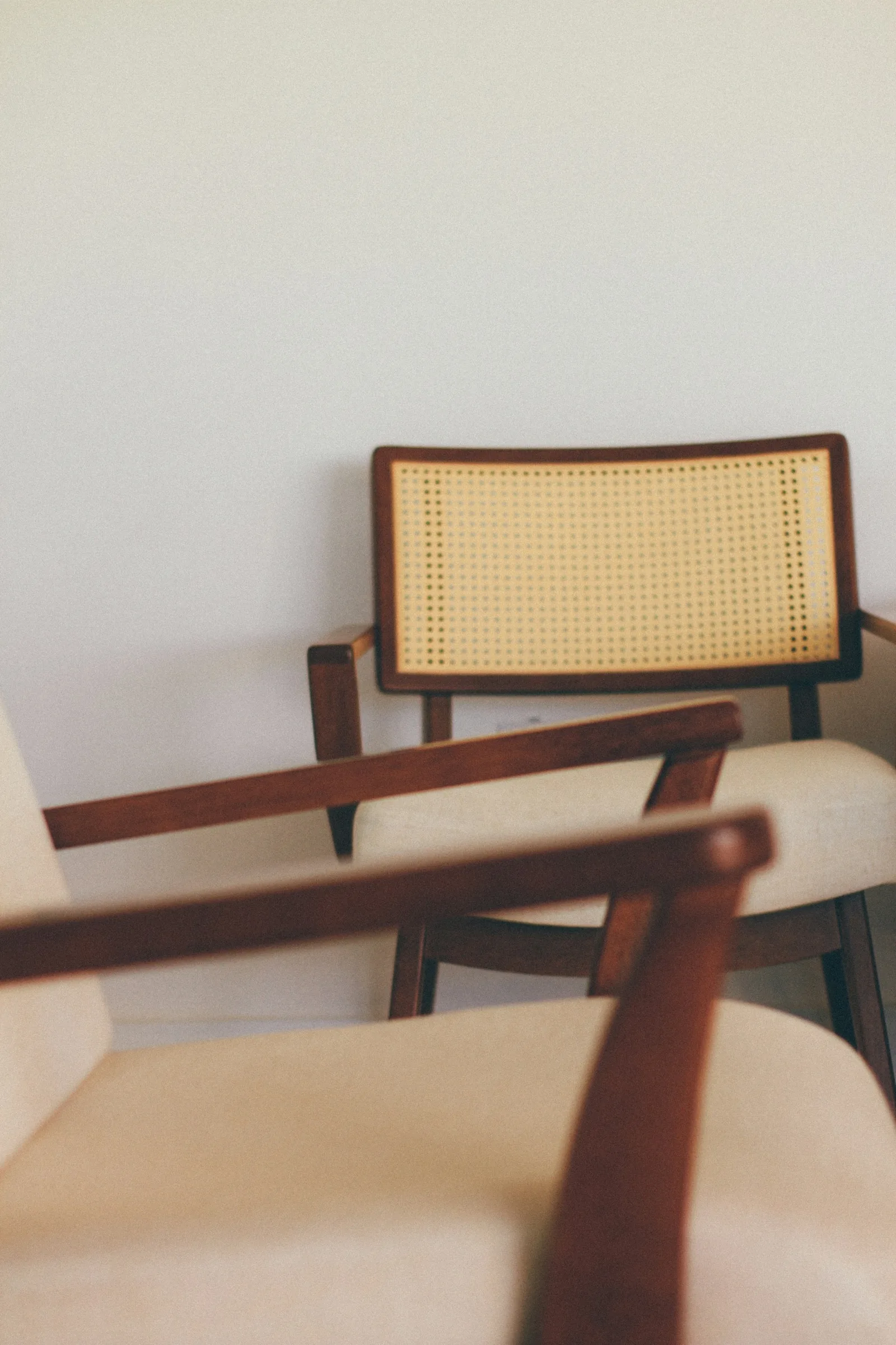 A low lounge chair with a cane-woven back, cushioned seat and long wooden armrests against a white wall