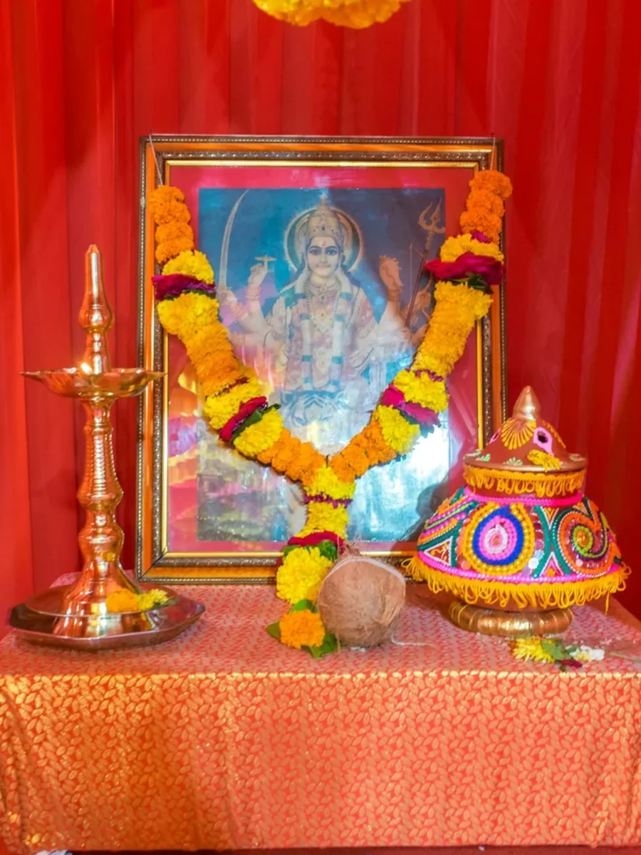A home shrine draped in orange-red fabric, with a garlanded deity picture, brass oil lamp and marigolds