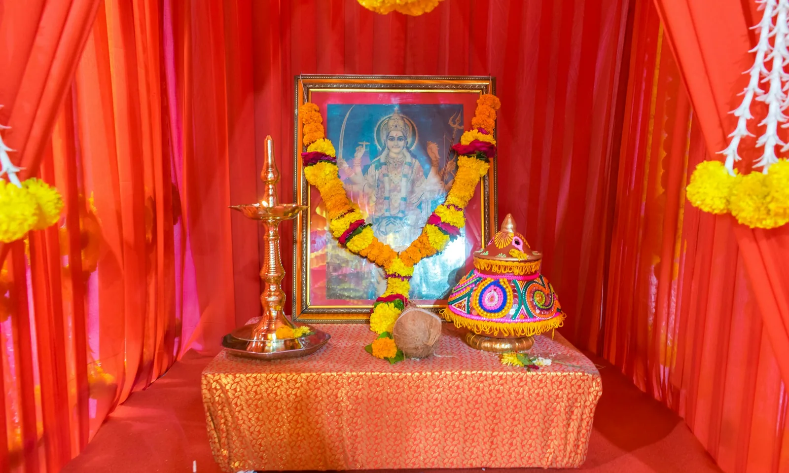 A home shrine draped in orange-red fabric, with a garlanded deity picture, brass oil lamp and marigolds