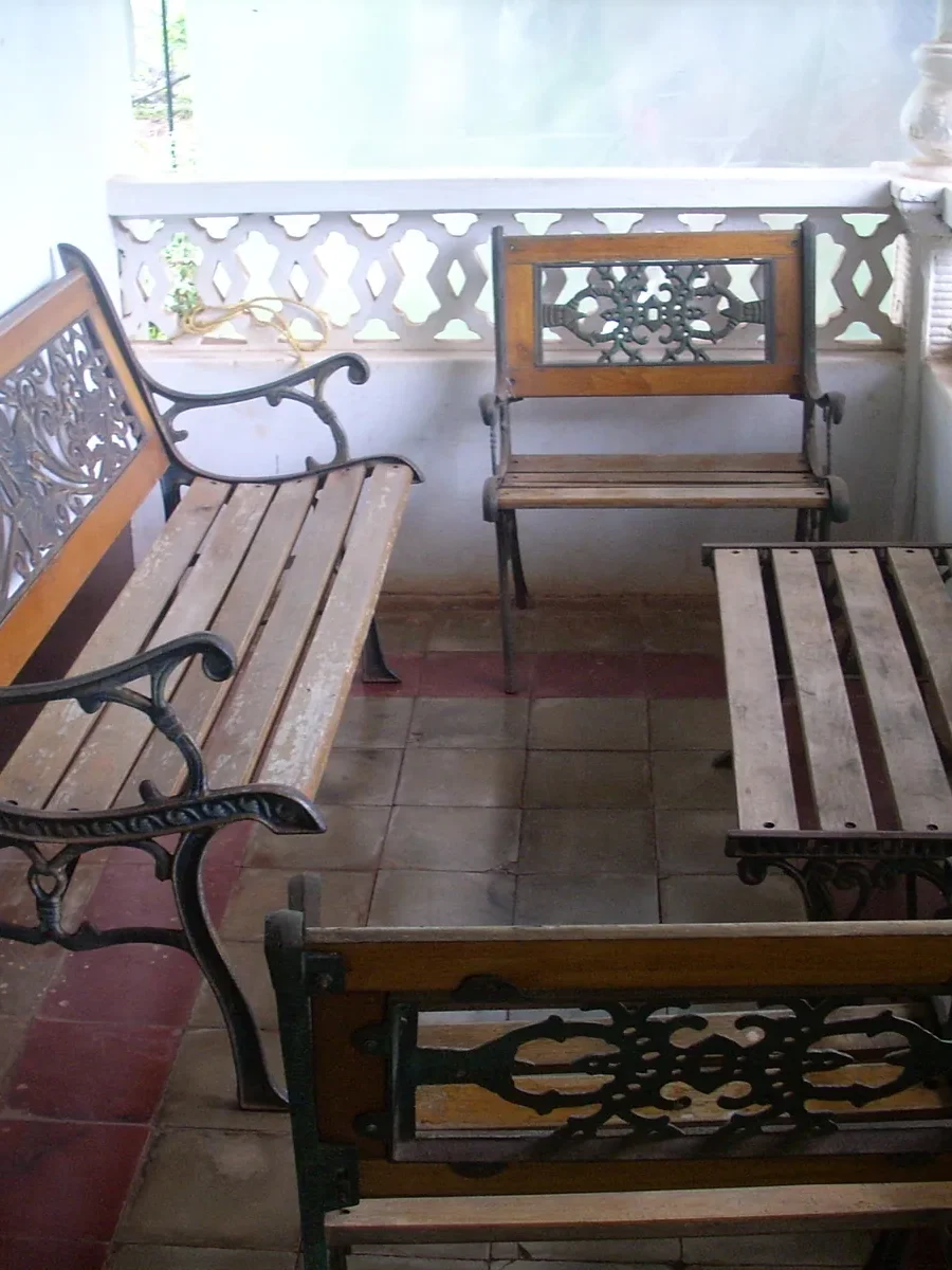 A shaded Goan verandah with cast-iron and timber benches against a pierced white balustrade