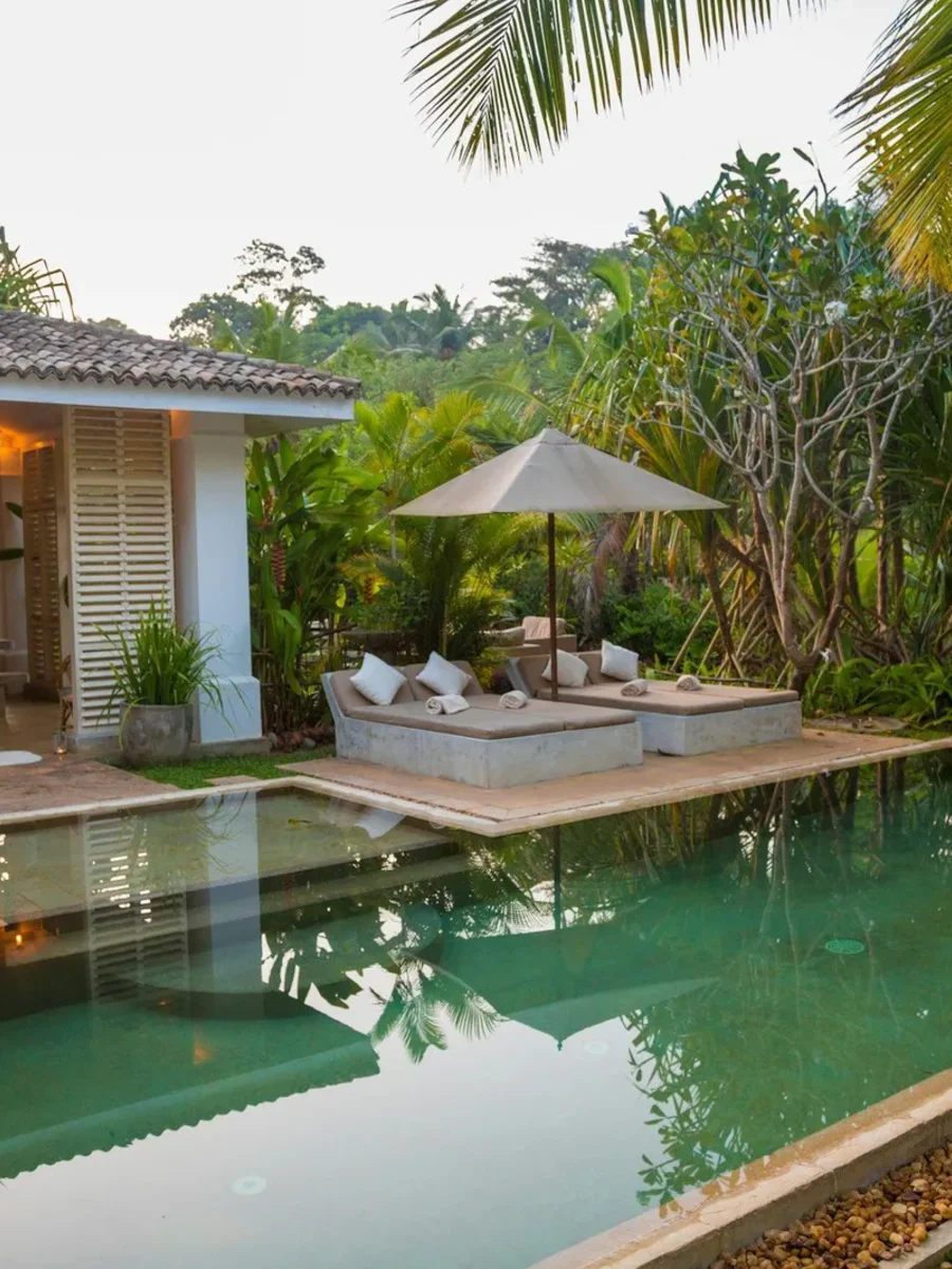 Green-tiled private pool beside a shuttered villa pavilion with daybeds, umbrella and palms