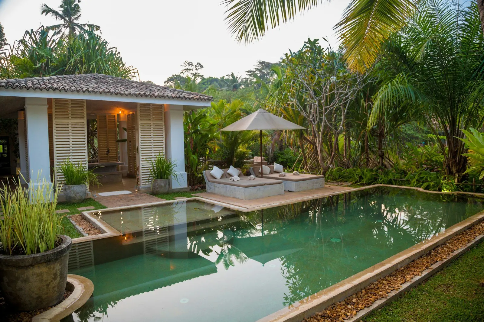 Green-tiled private pool beside a shuttered villa pavilion with daybeds, umbrella and palms