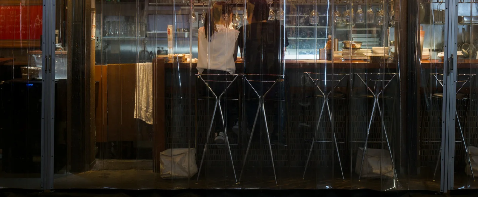 Clear PVC curtain panels enclosing a restaurant counter, with two diners seen from behind on bar stools