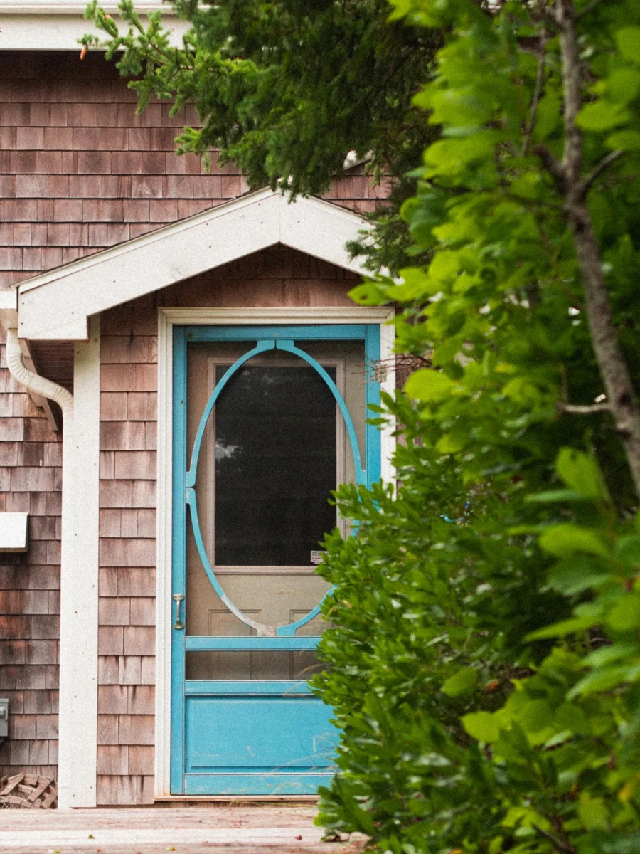 A turquoise wooden screen door with an oval mesh panel at the entrance of a shingled house