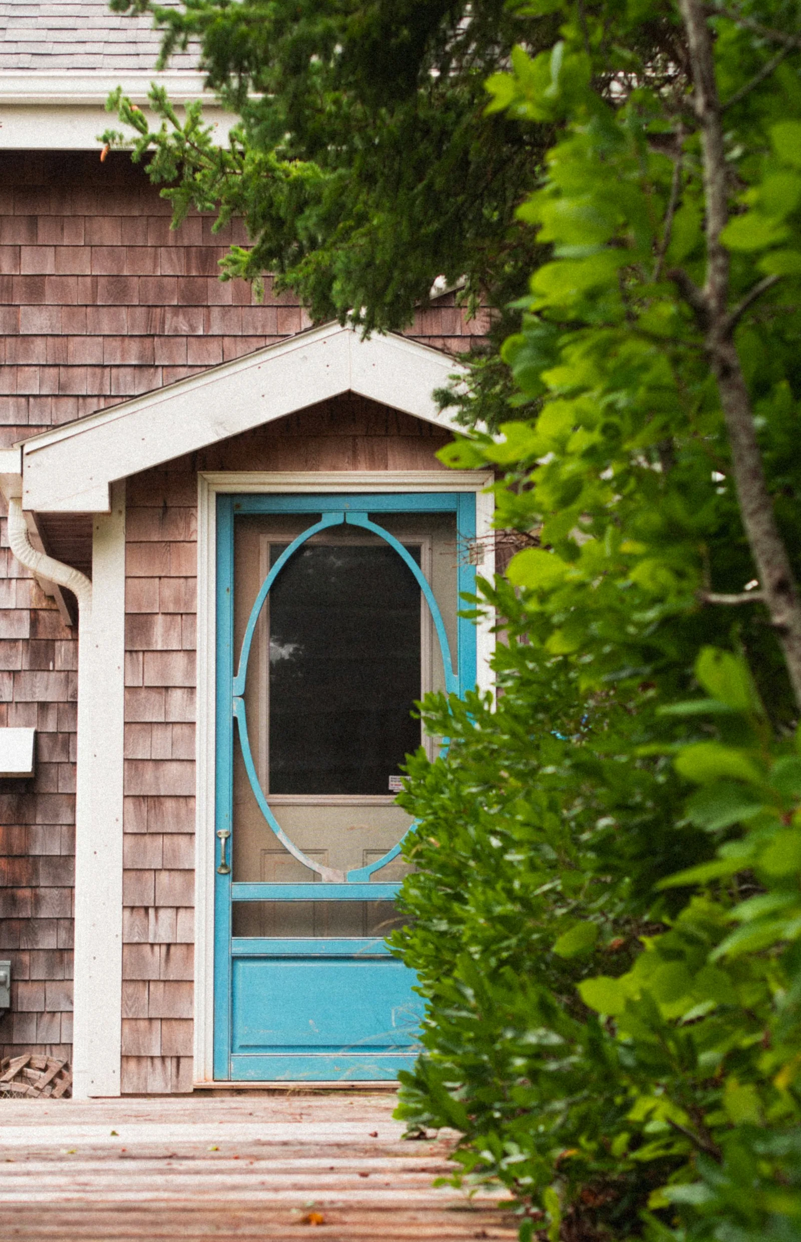 A turquoise wooden screen door with an oval mesh panel at the entrance of a shingled house