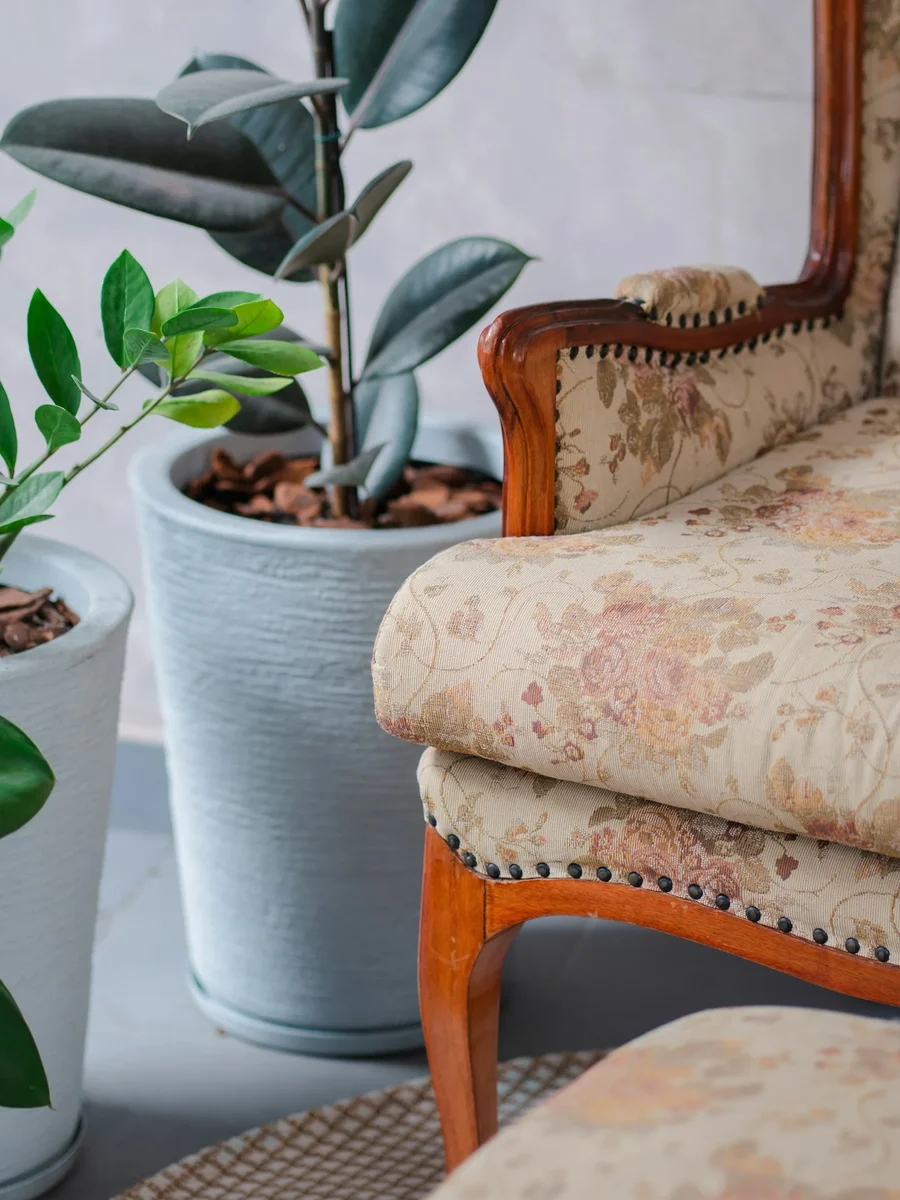An antique carved-wood armchair with worn floral upholstery and studded trim beside potted plants