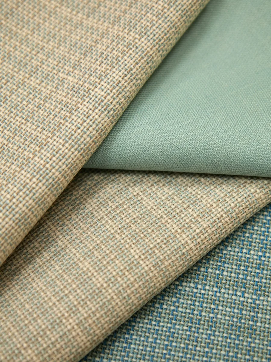 Folded upholstery fabric swatches in oatmeal tweed, sage green and teal weaves laid over each other