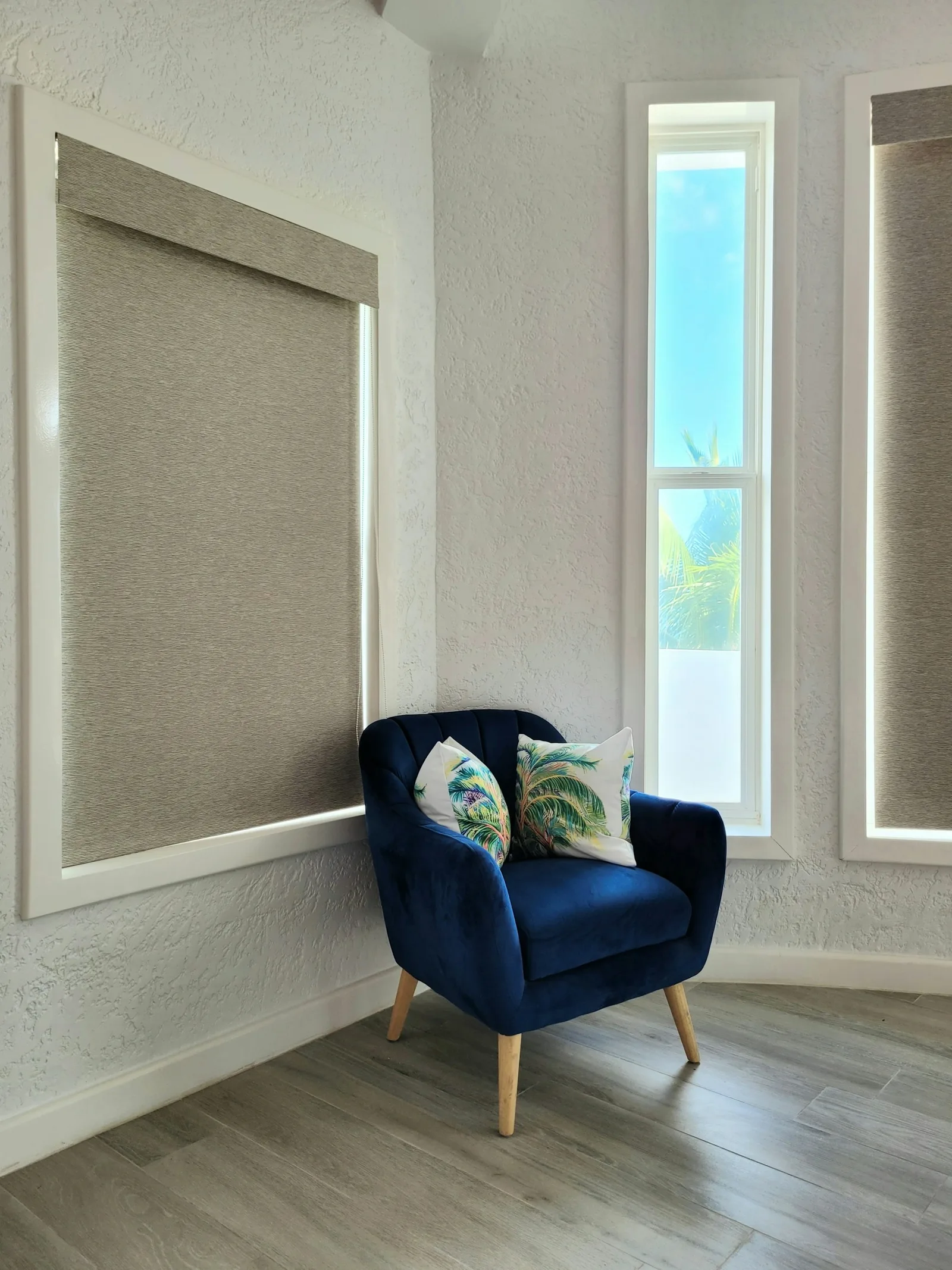 A grey fabric roller blind lowered over a window beside a blue velvet armchair