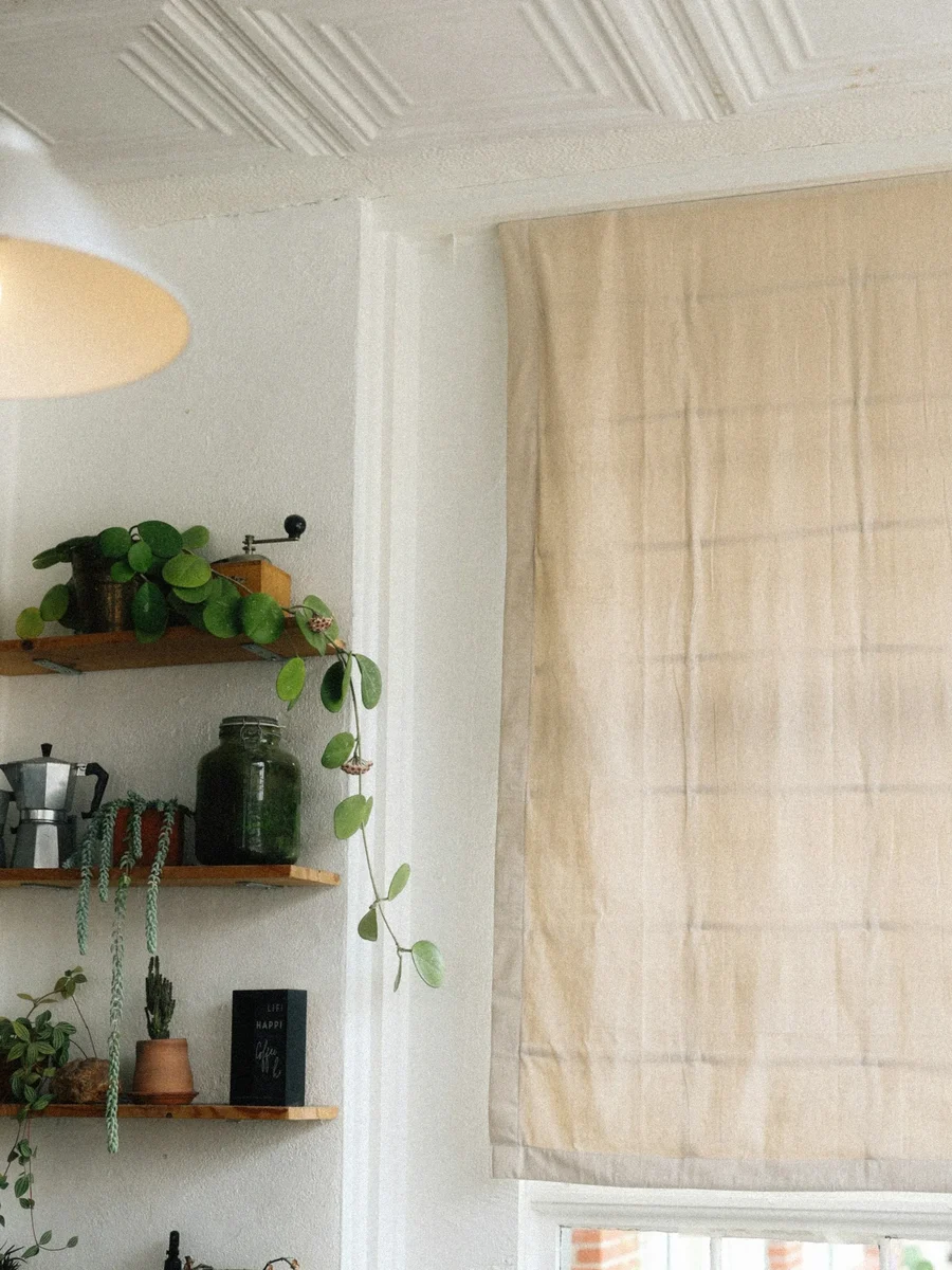A linen Roman blind lowered over a tall window beside open shelves of plants and coffee things