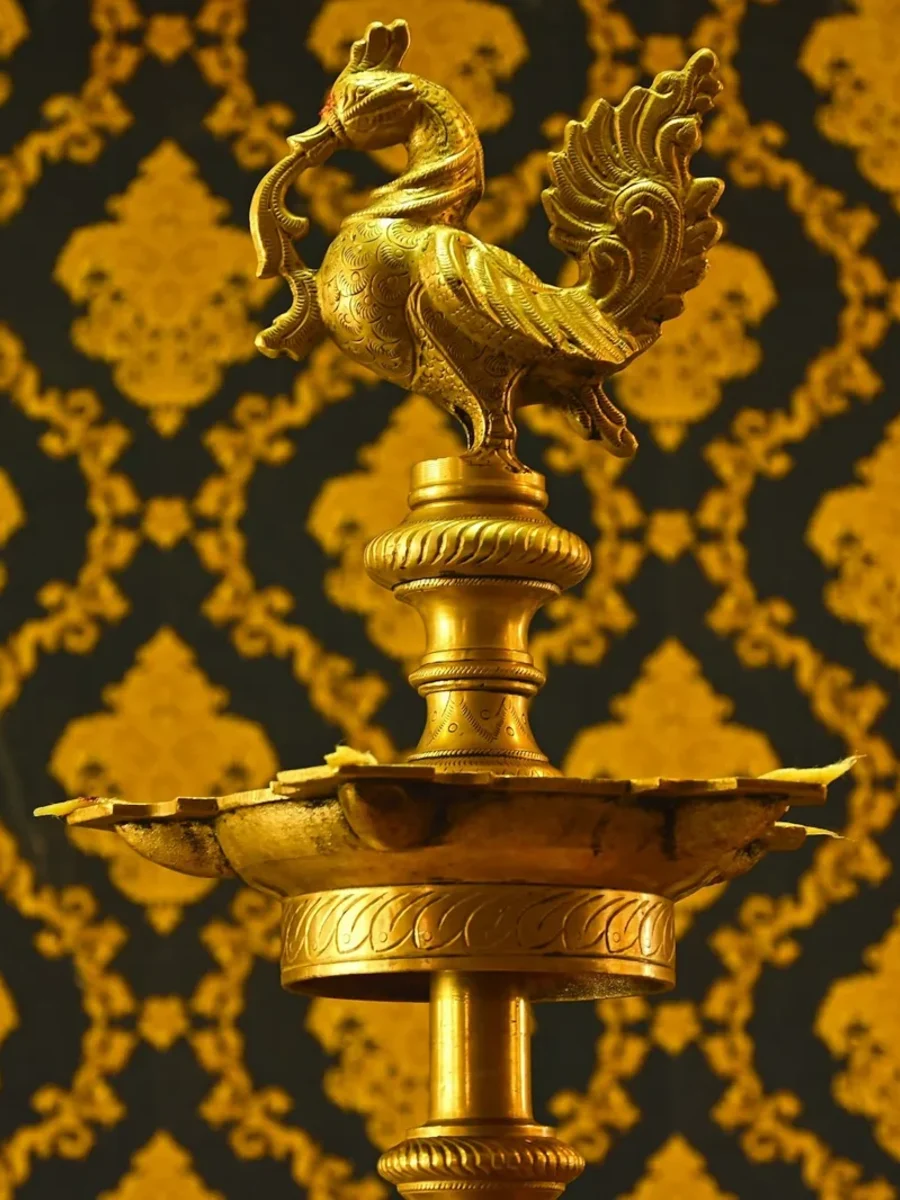A tall brass oil lamp with a peacock finial and petal-shaped wick tray against gold-patterned wallpaper