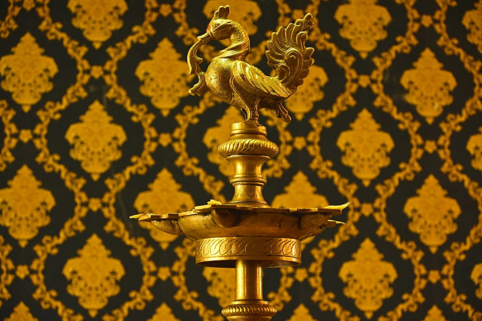 A tall brass oil lamp with a peacock finial and petal-shaped wick tray against gold-patterned wallpaper