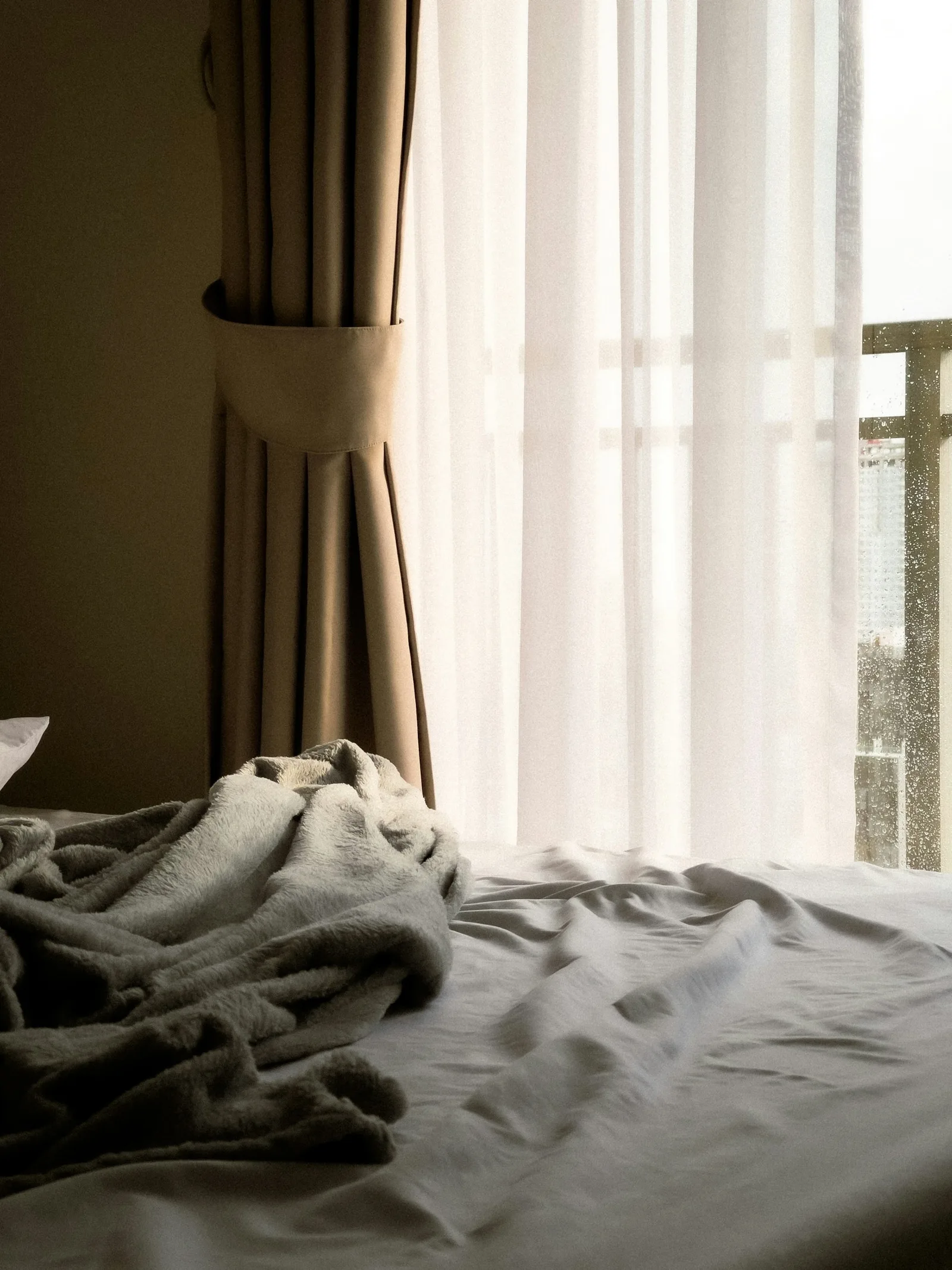 A taupe curtain held back with a tie beside a white sheer, over an unmade bed in morning light