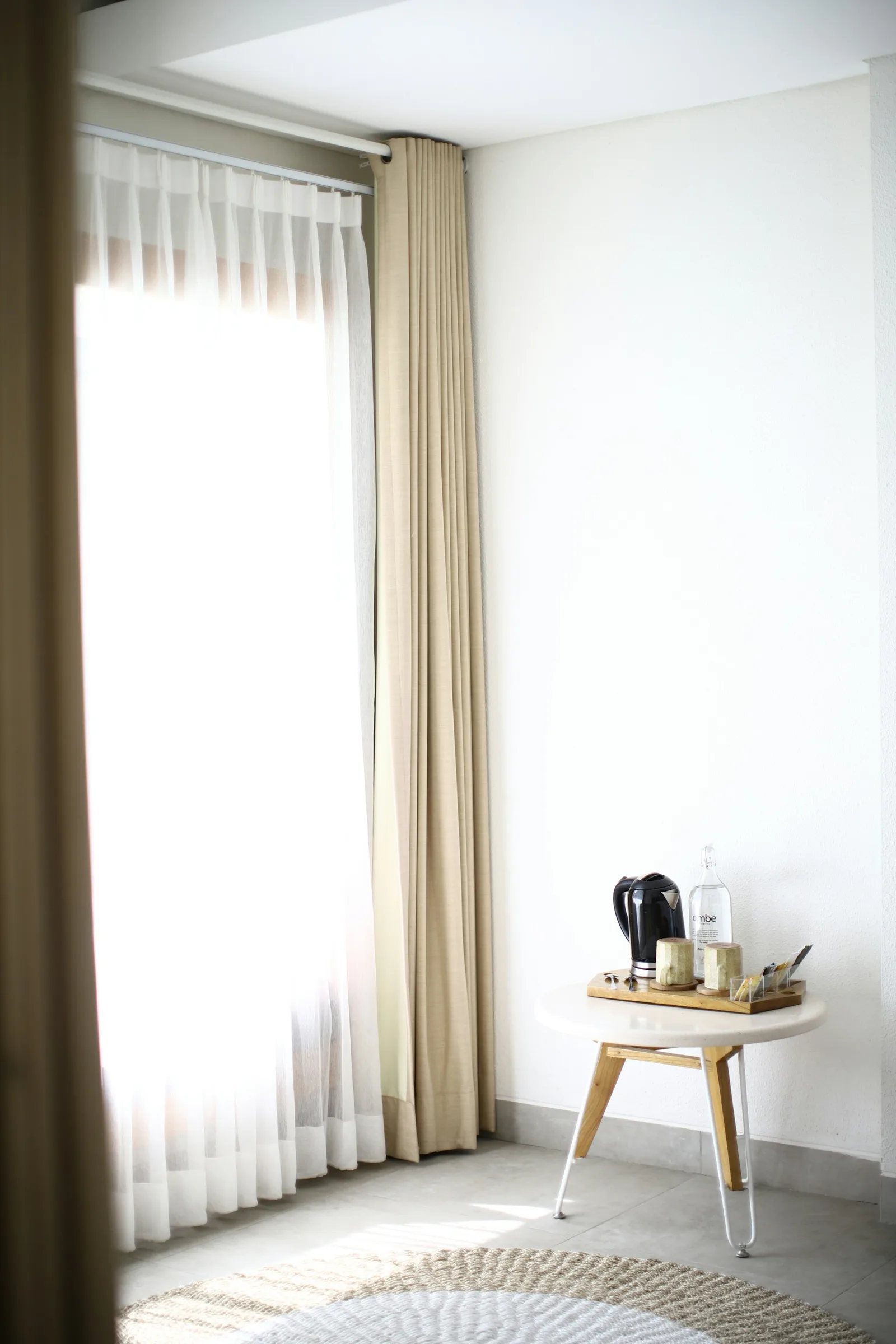 A tall window dressed with a white sheer and a heavier beige side curtain, beside a small side table