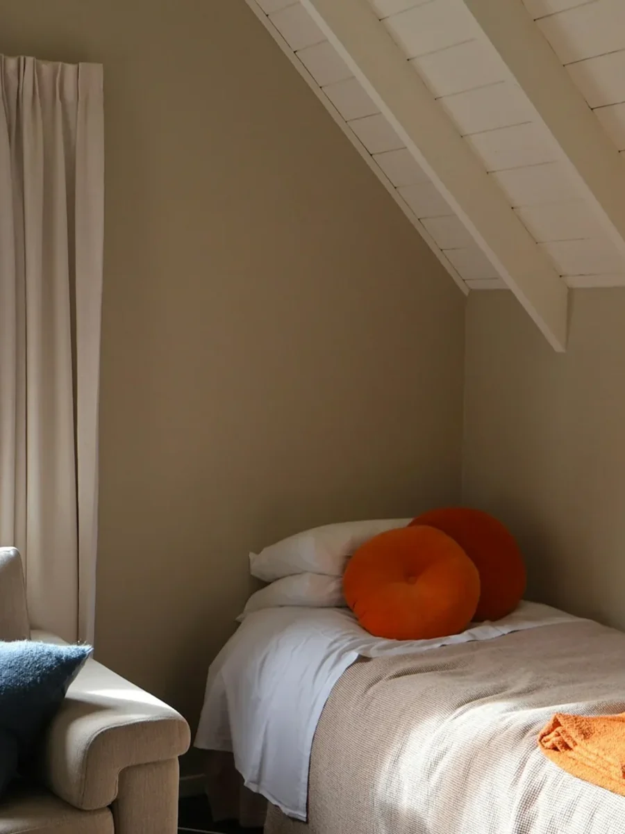 A compact attic bedroom with a single bed, orange cushions and linen curtains at a garden window