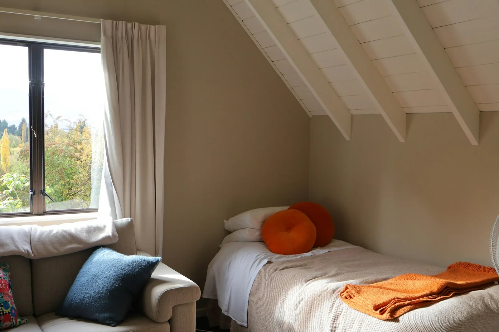 A compact attic bedroom with a single bed, orange cushions and linen curtains at a garden window