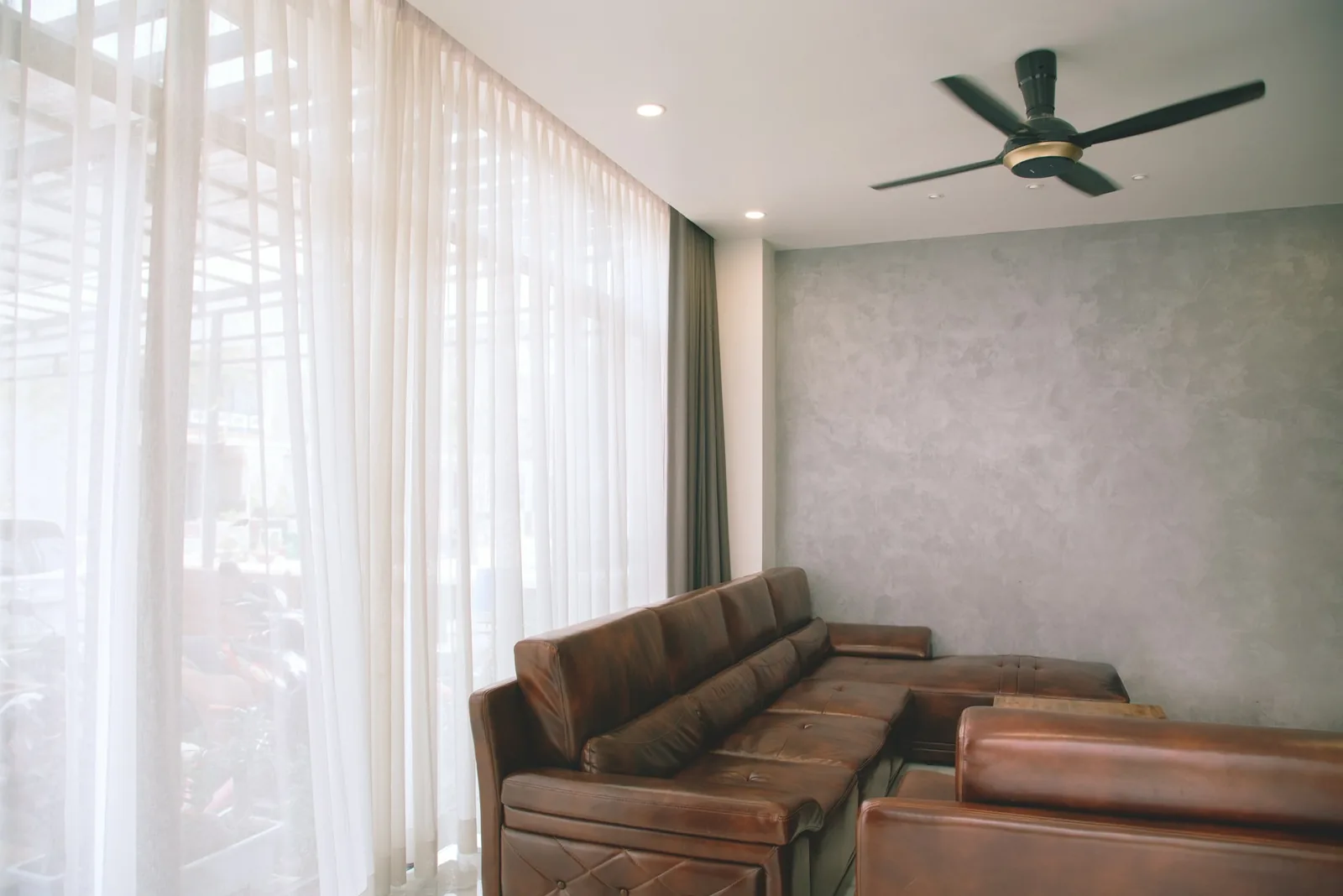 Floor-to-ceiling sheer curtains on a ceiling track beside a leather sofa and ceiling fan