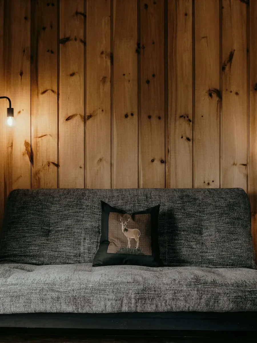 Grey futon-style sofa bed against a knotty pine wall, with a floor lamp and log side table
