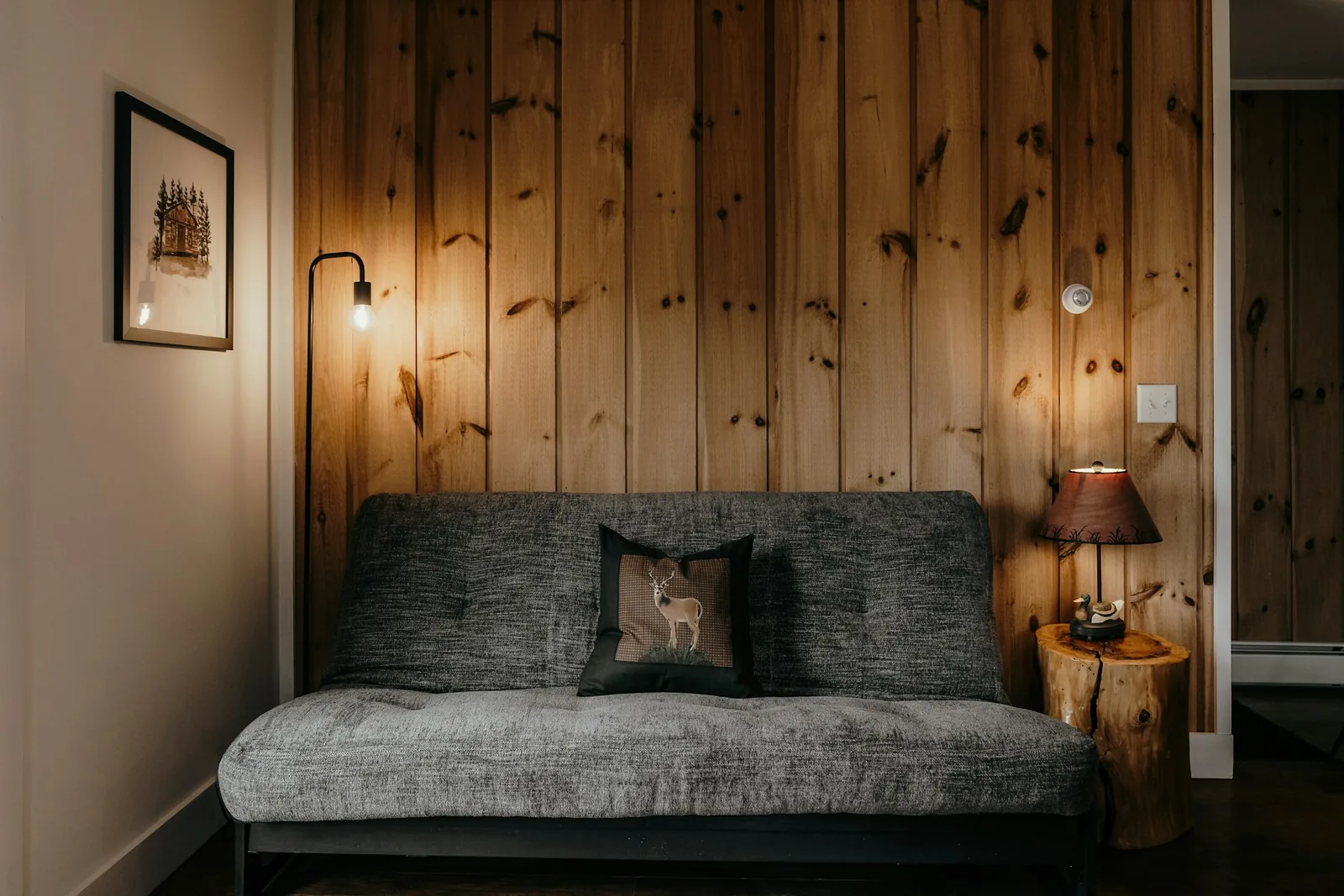 Grey futon-style sofa bed against a knotty pine wall, with a floor lamp and log side table