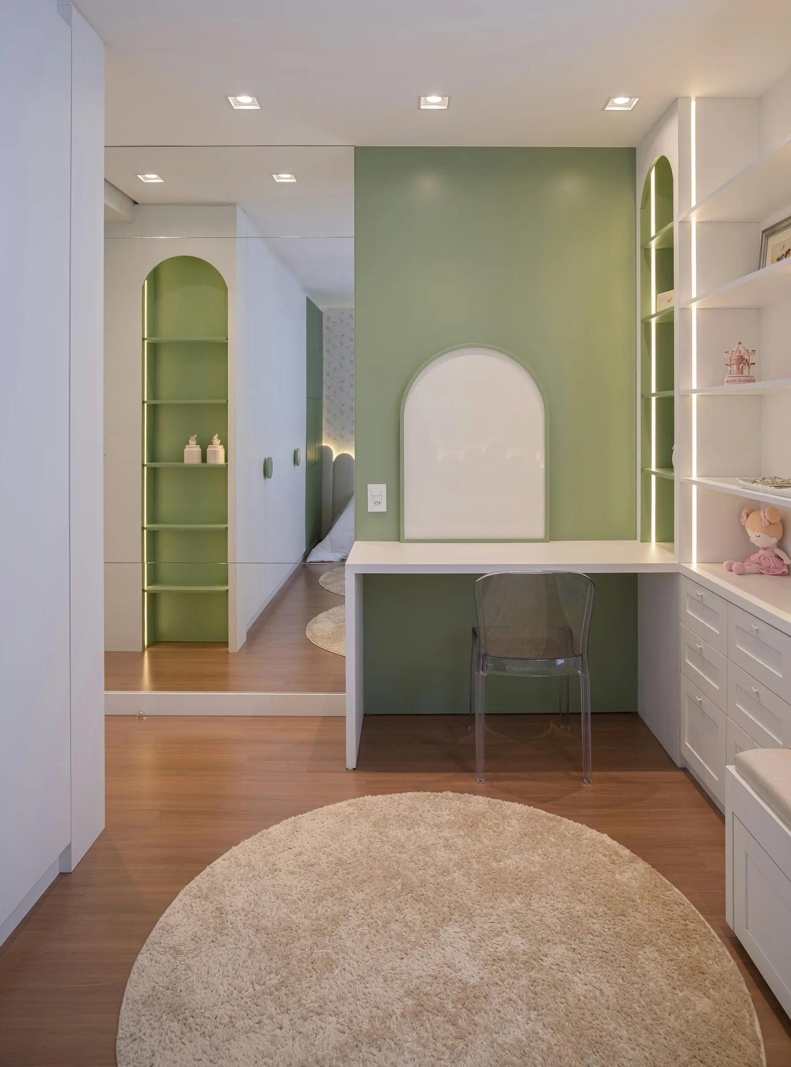 A study nook with a built-in white desk, sage-green wall, arched shelves and a round rug