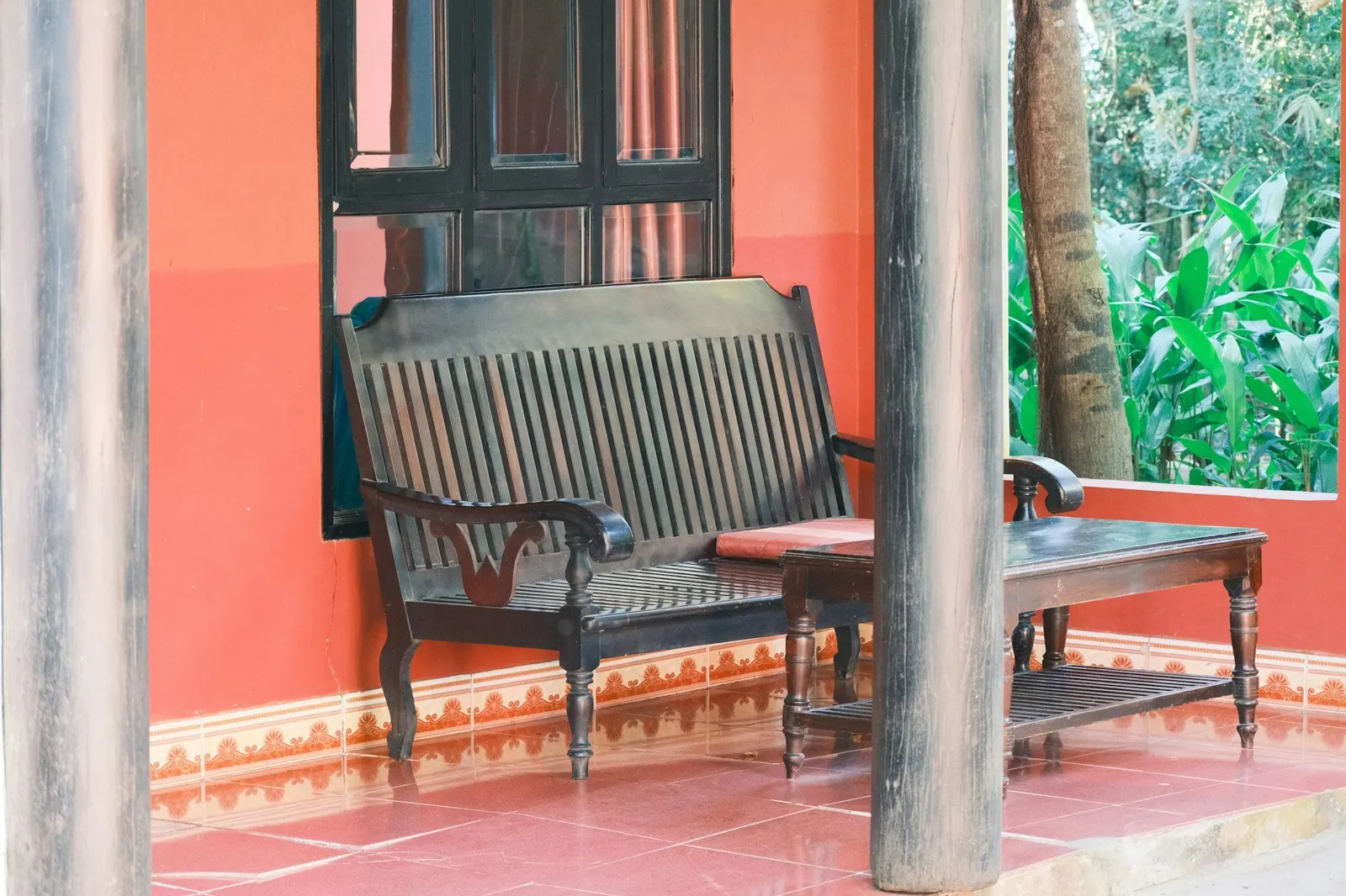 Dark wooden bench and table on a red-tiled verandah between timber pillars and greenery