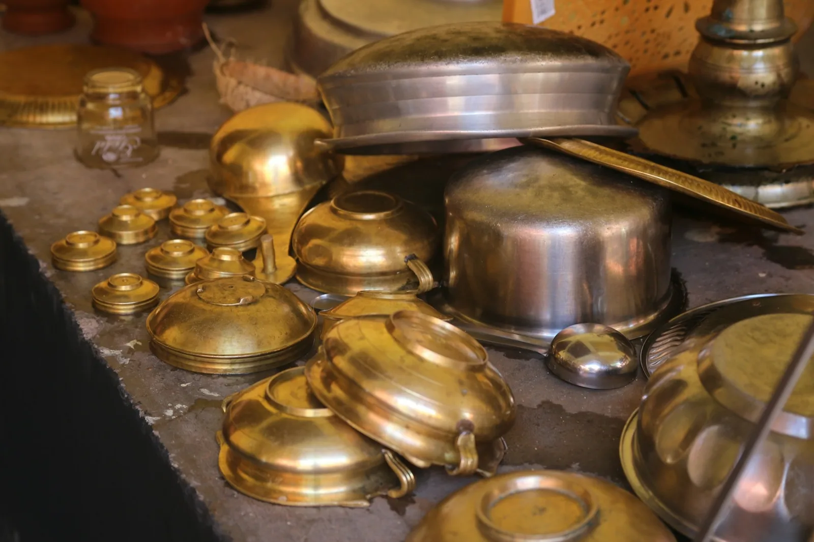 Brass bowls, lids and steel cooking pots heaped on a stone counter in an Indian kitchen