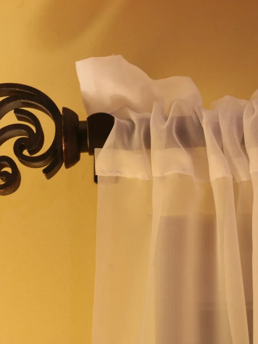 Wrought-iron curtain rod with a scrolled finial holding a white sheer curtain on a warm wall