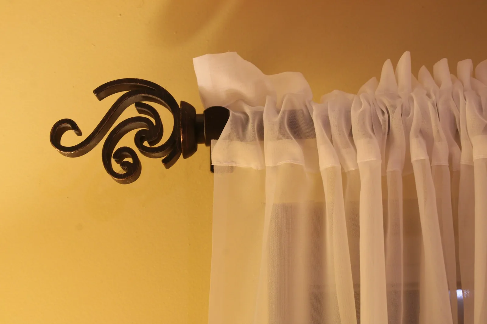 Wrought-iron curtain rod with a scrolled finial holding a white sheer curtain on a warm wall
