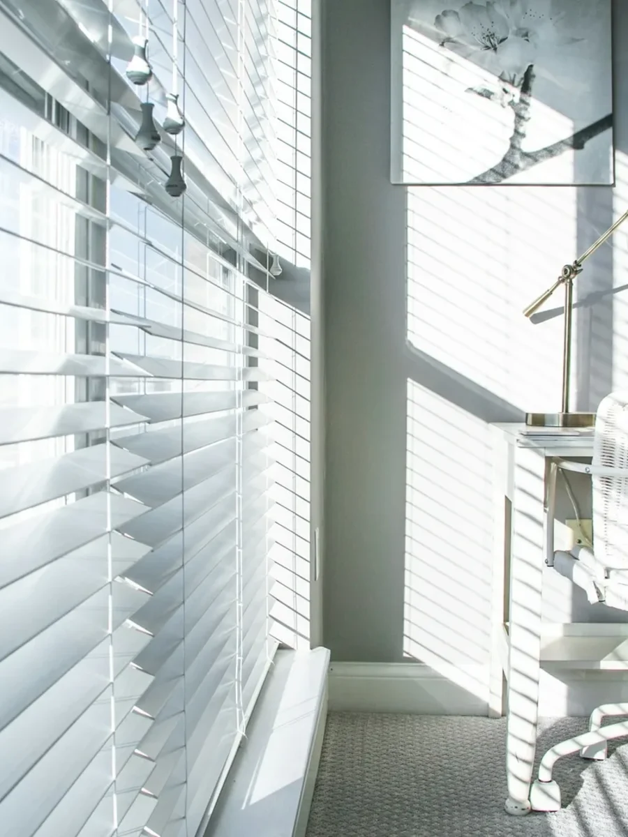 White venetian blinds tilted to stripe sunlight across a small home office desk and chair