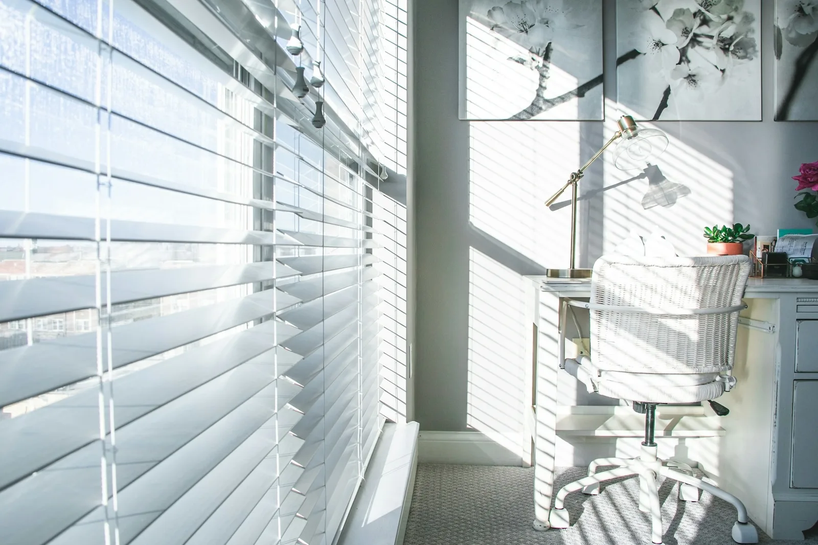 White venetian blinds tilted to stripe sunlight across a small home office desk and chair