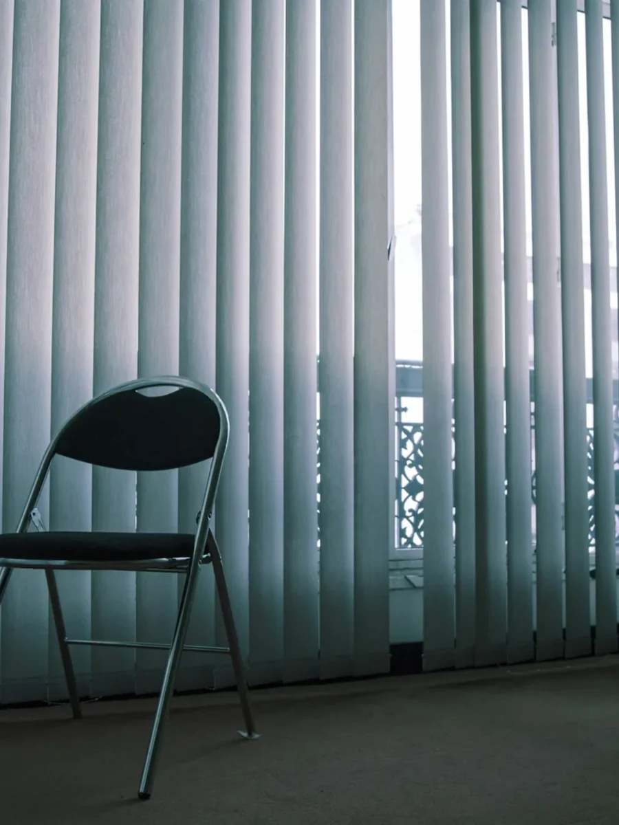 Pale fabric vertical blind slats across a tall window, with a folding chair in front