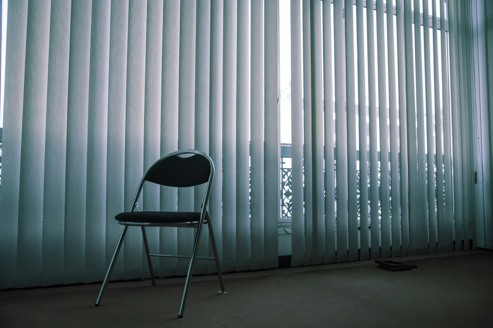Pale fabric vertical blind slats across a tall window, with a folding chair in front