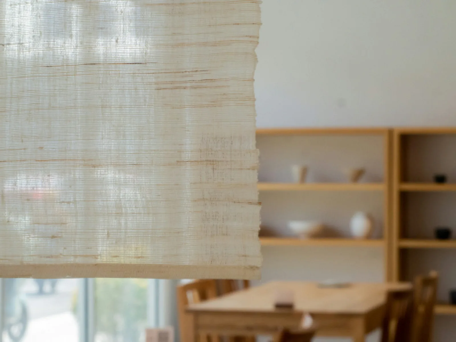 Close view of a raw-linen fabric blind lowered at a window, wooden shelves blurred behind