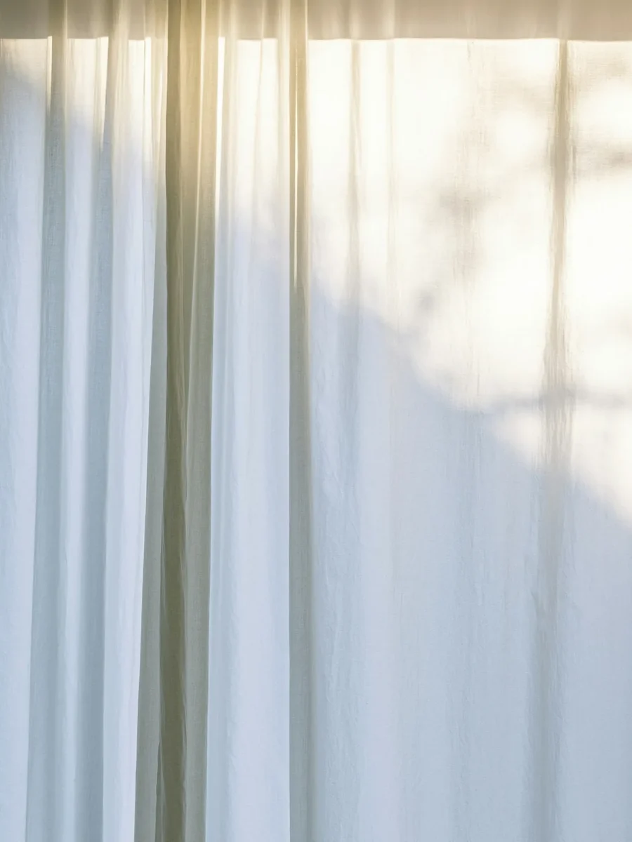 Soft white cotton curtains catching warm sunlight and the shadow of leaves outside the window